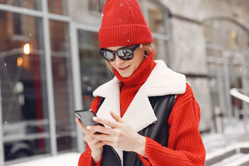 Une femme en tuque rouge consulte son téléphone dans une rue enneigée.