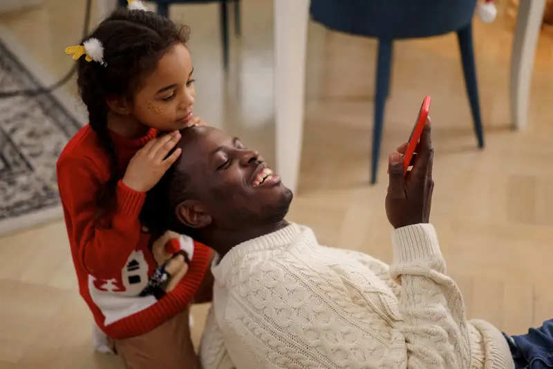 Un père allongé sur le plancher sourit, téléphone en main, pendant que sa fille en chandail des fêtes l'enlace.