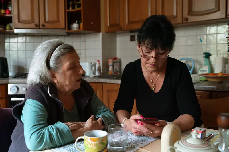 Une femme et sa mère âgée discutent à la table de cuisine, un téléphone à la main.